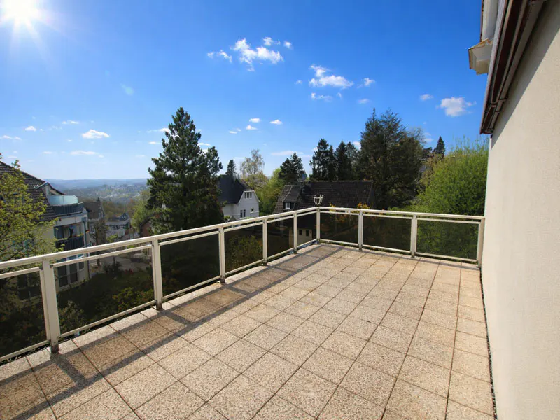 Tiled balcony with glass railings overlooks a neighborhood with trees under a blue sky with clouds and a bright sun.