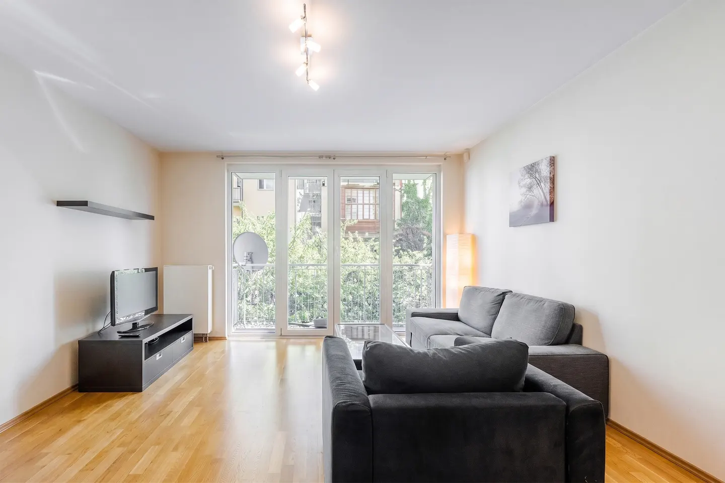 Living room with wood floors, white walls, and gray sofas. Balcony doors show greenery outside. A TV sits on a black stand.