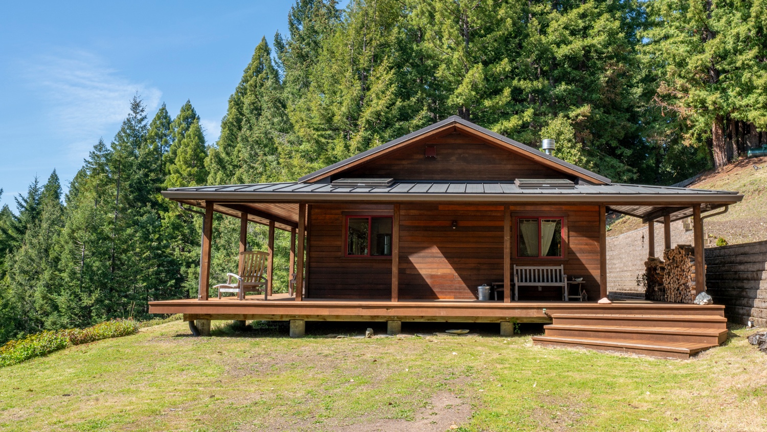 A rustic wooden cabin with a wrap-around porch, outdoor seating, and a woodpile, nestled in a lush forest setting.