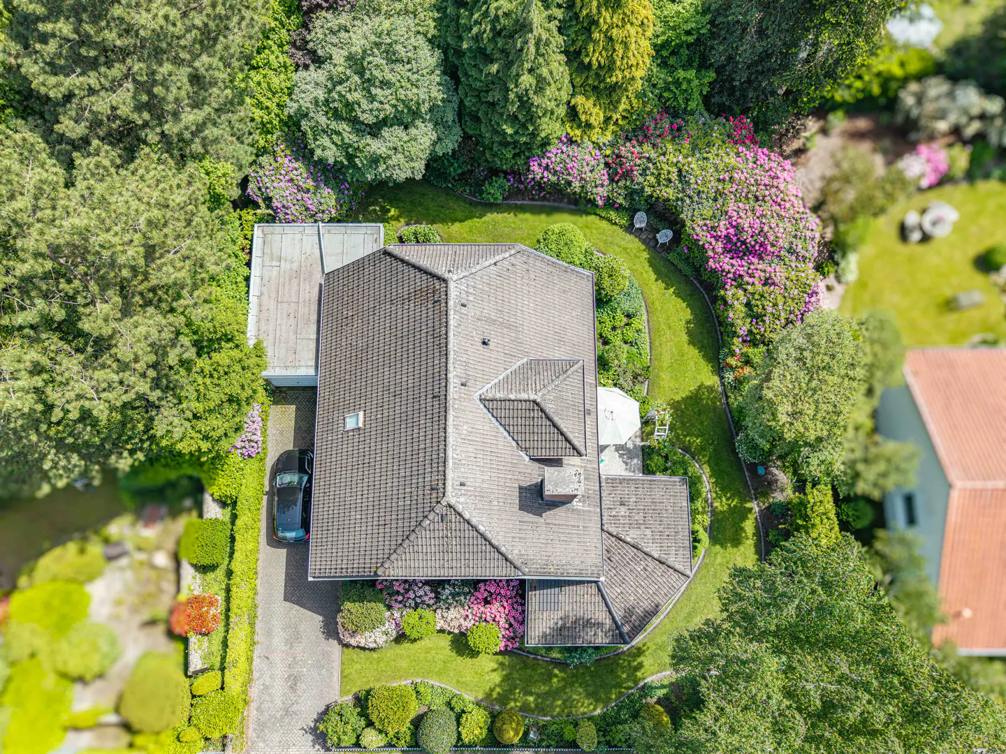 Aerial view of a house with a gray roof, surrounded by lush green trees and pink flowers. A car is parked in the driveway.