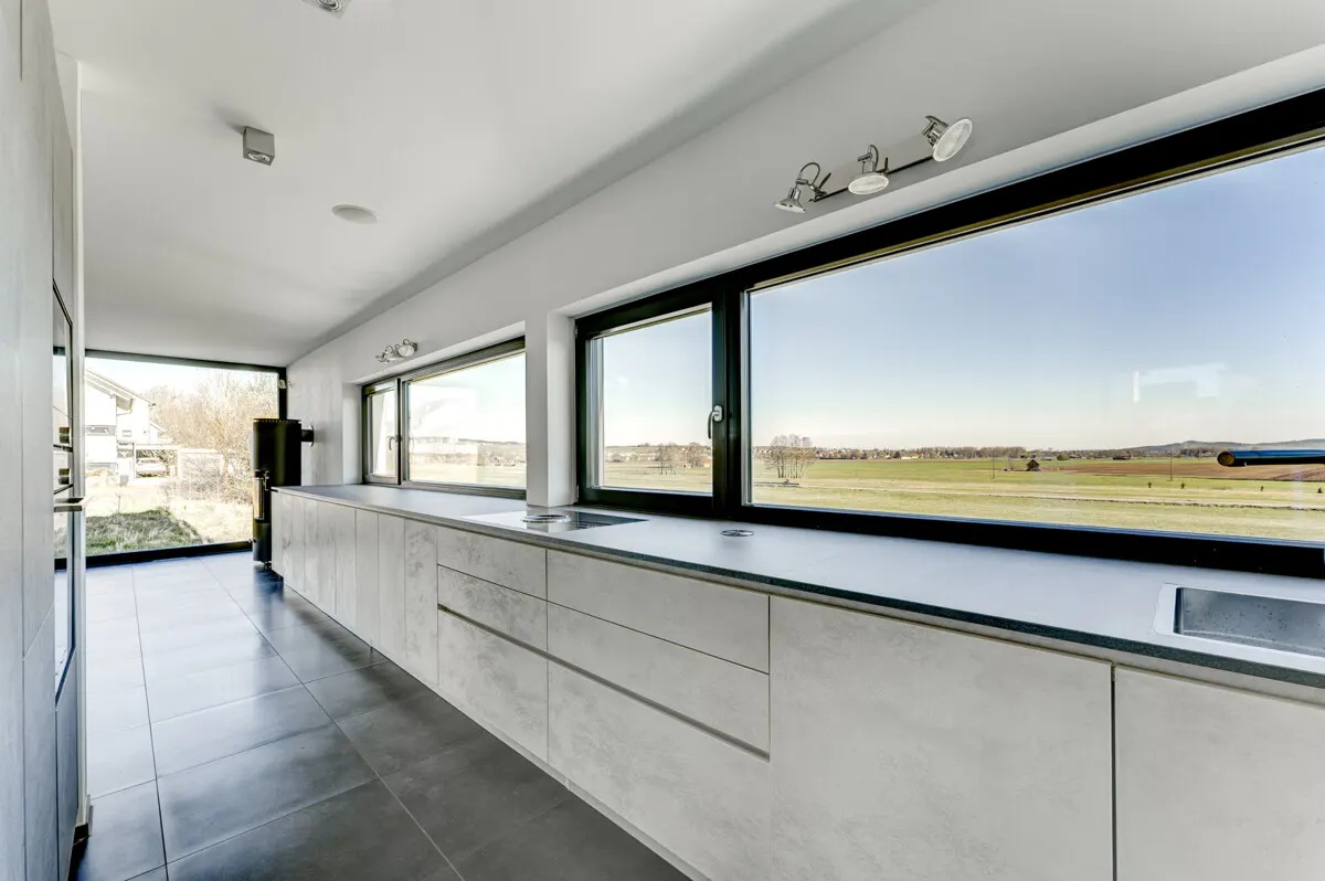 A modern kitchen with gray cabinets, black framed windows, and gray tile floors. Outside the windows is a green field.