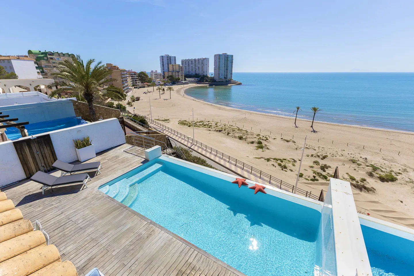 Rooftop pool with two starfish floats, wooden deck, and lounge chairs overlooking a sandy beach and blue ocean on a sunny day.