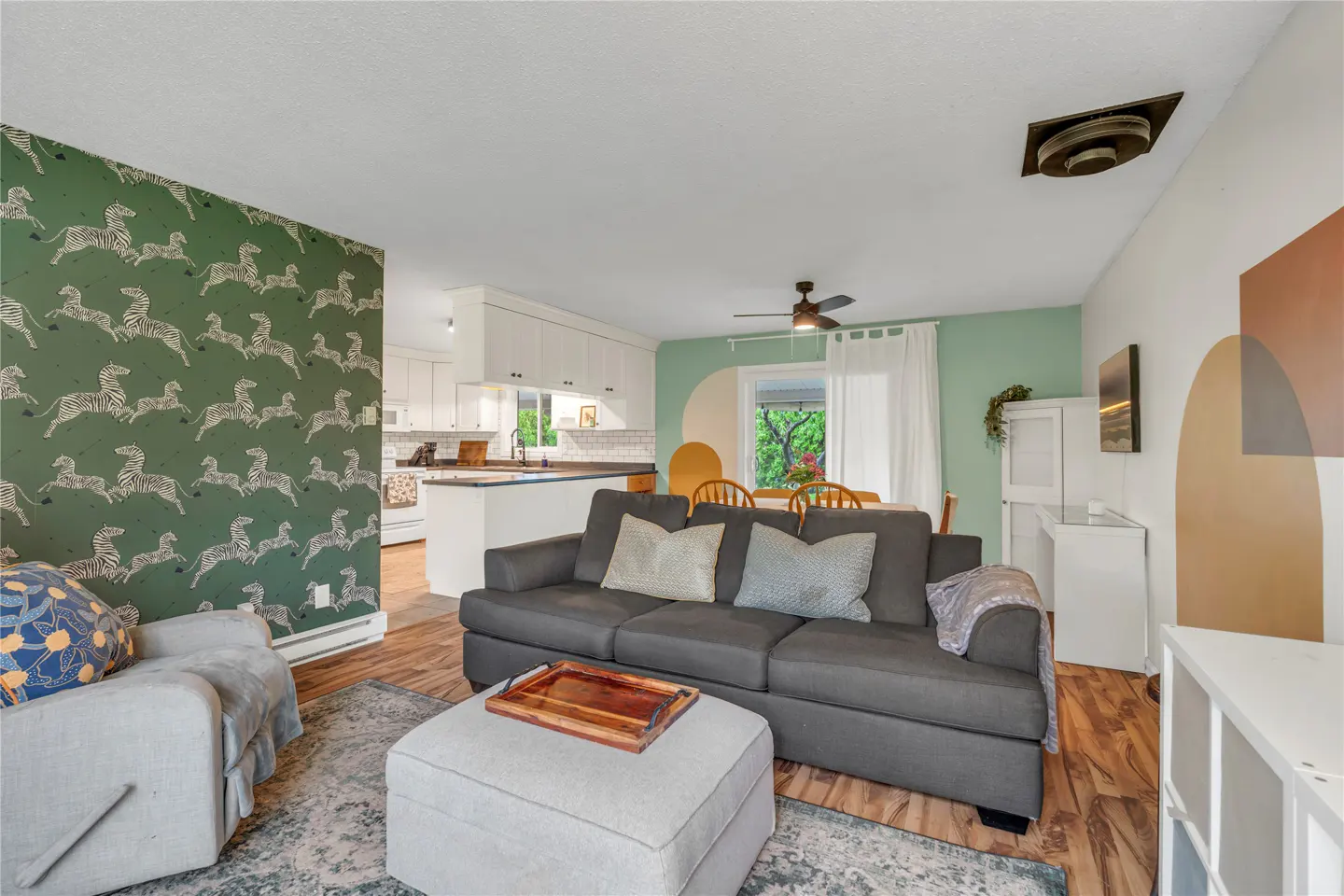 Living room with dark gray sofa, light gray ottoman, zebra-patterned green wallpaper, and open kitchen in the background.