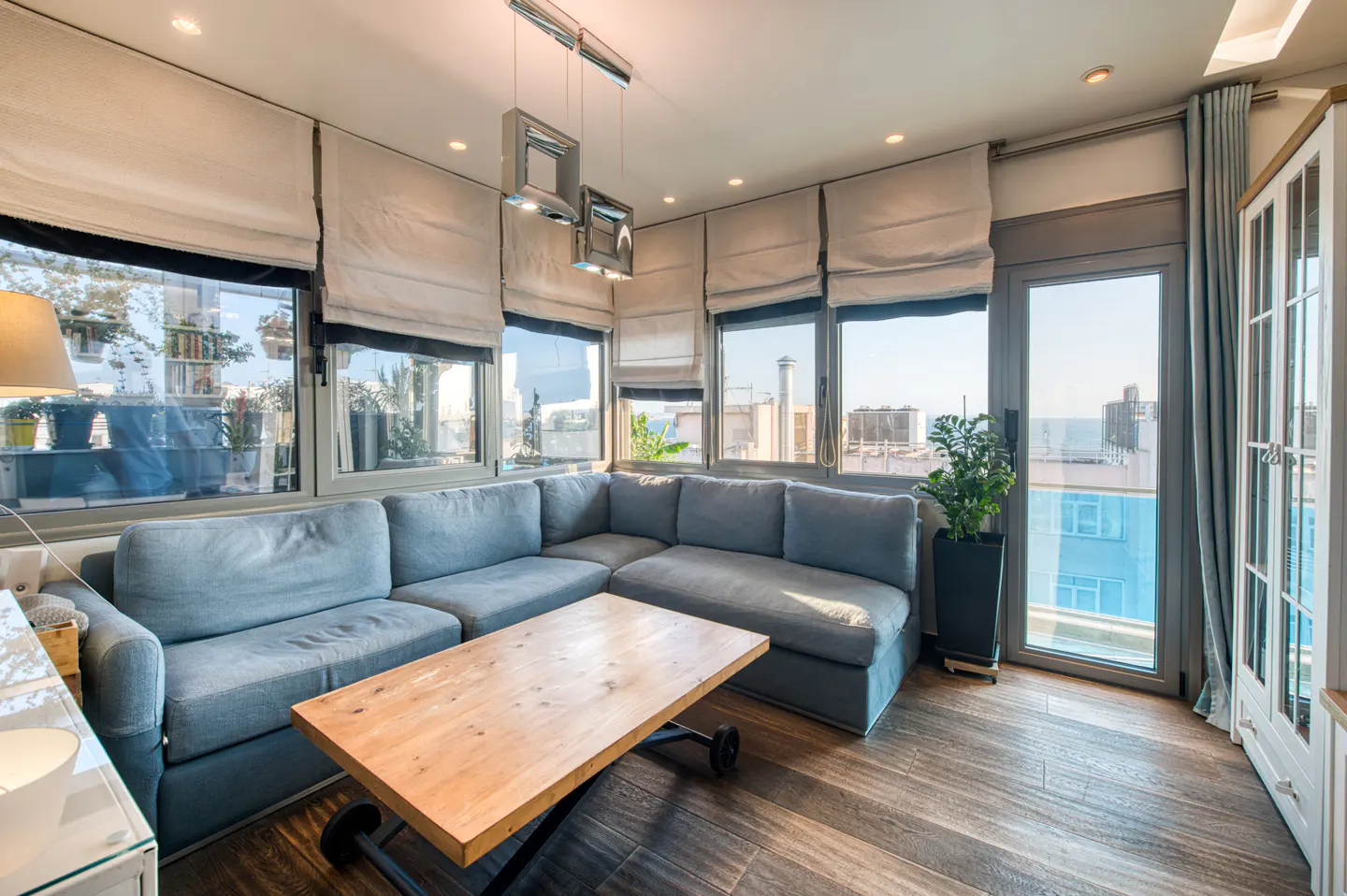 Bright living room with a blue sectional sofa, wood coffee table, and large windows with white blinds overlooking a cityscape.