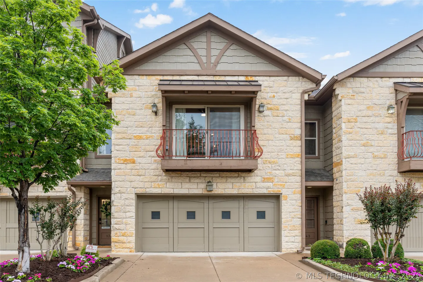 Exterior of a two-story townhome with a beige stone facade, a brown roof, and a two-car garage. A small red balcony is above the garage.