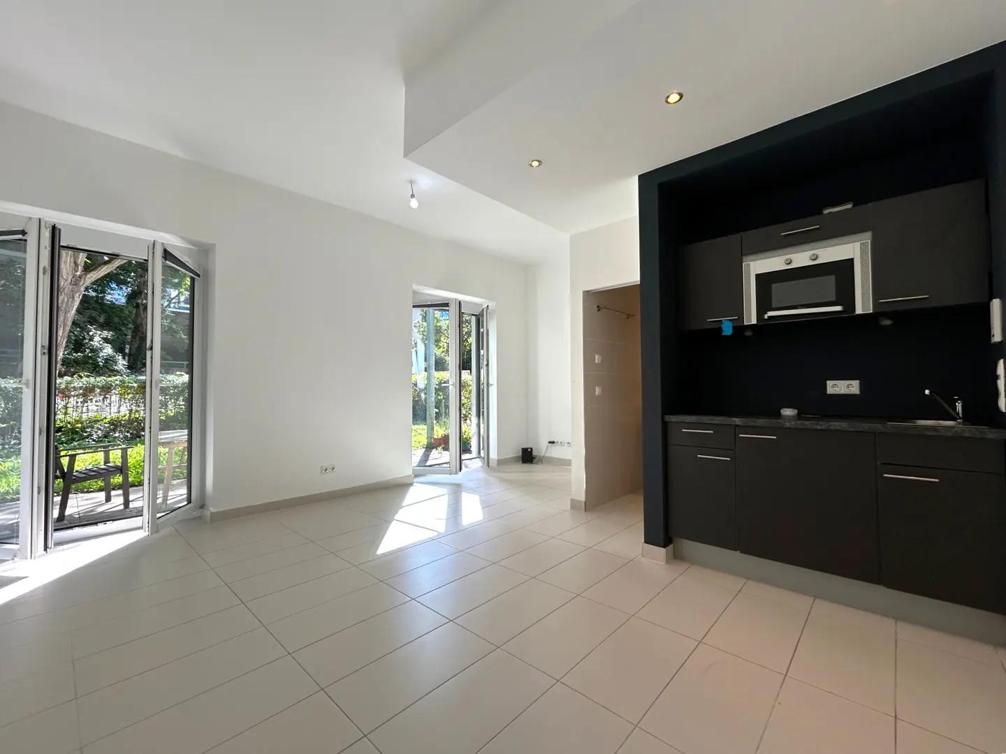 Bright, empty apartment with white tile floors, open patio doors, and a dark kitchenette with microwave.