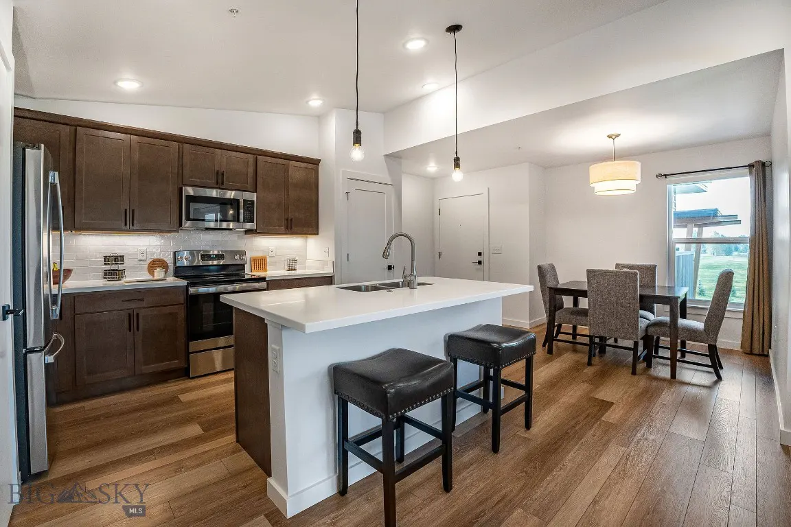 A modern kitchen with brown cabinets, stainless steel appliances, and a white island with black stools. A dining table sits near a window.