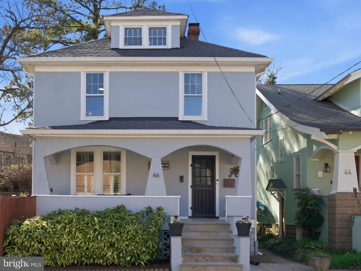 Two-story light blue house with a black door and roof, white trim, and green bushes in front.