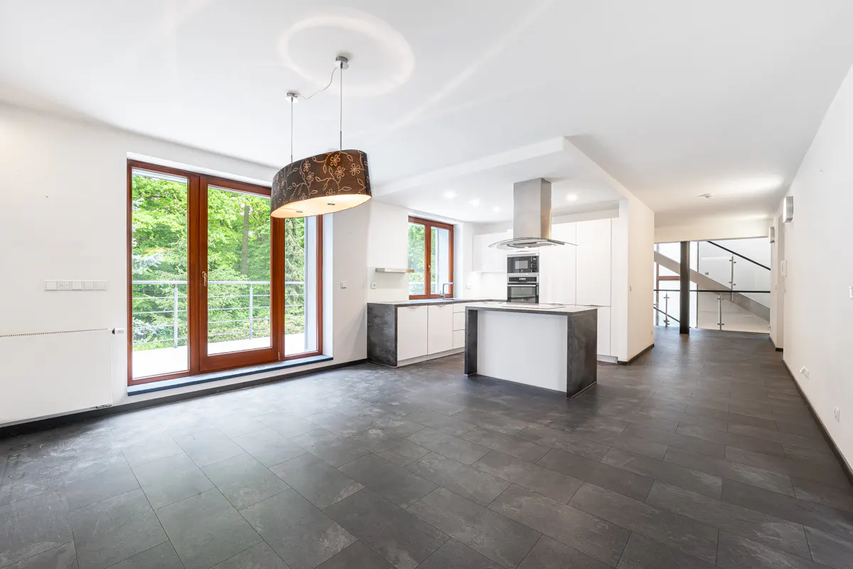 Bright, open-plan kitchen with dark tile floor, white cabinets, island, and wood-framed windows overlooking green trees.