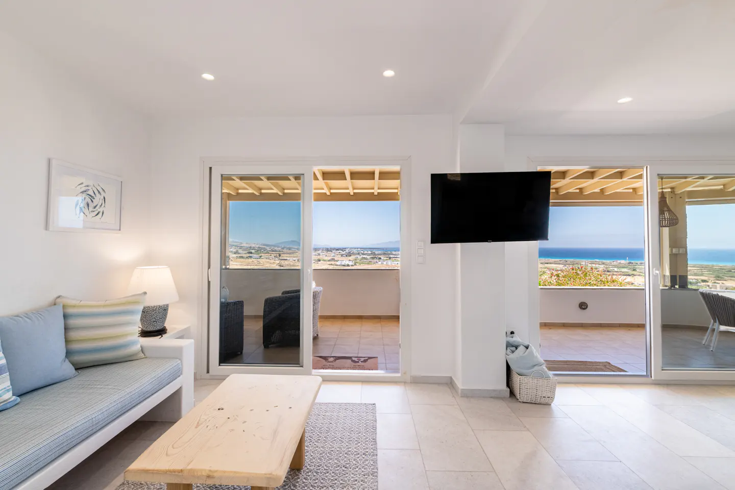 Bright living room with a sofa, wooden coffee table, wall-mounted TV, and sliding doors opening to a scenic ocean view.