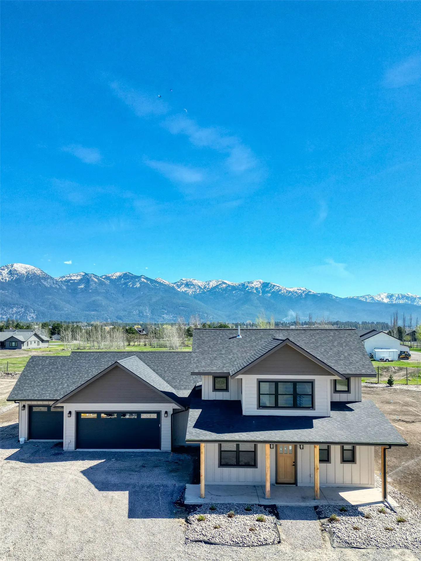 A modern, light-colored house with a dark roof and a mountain range in the background under a clear blue sky.