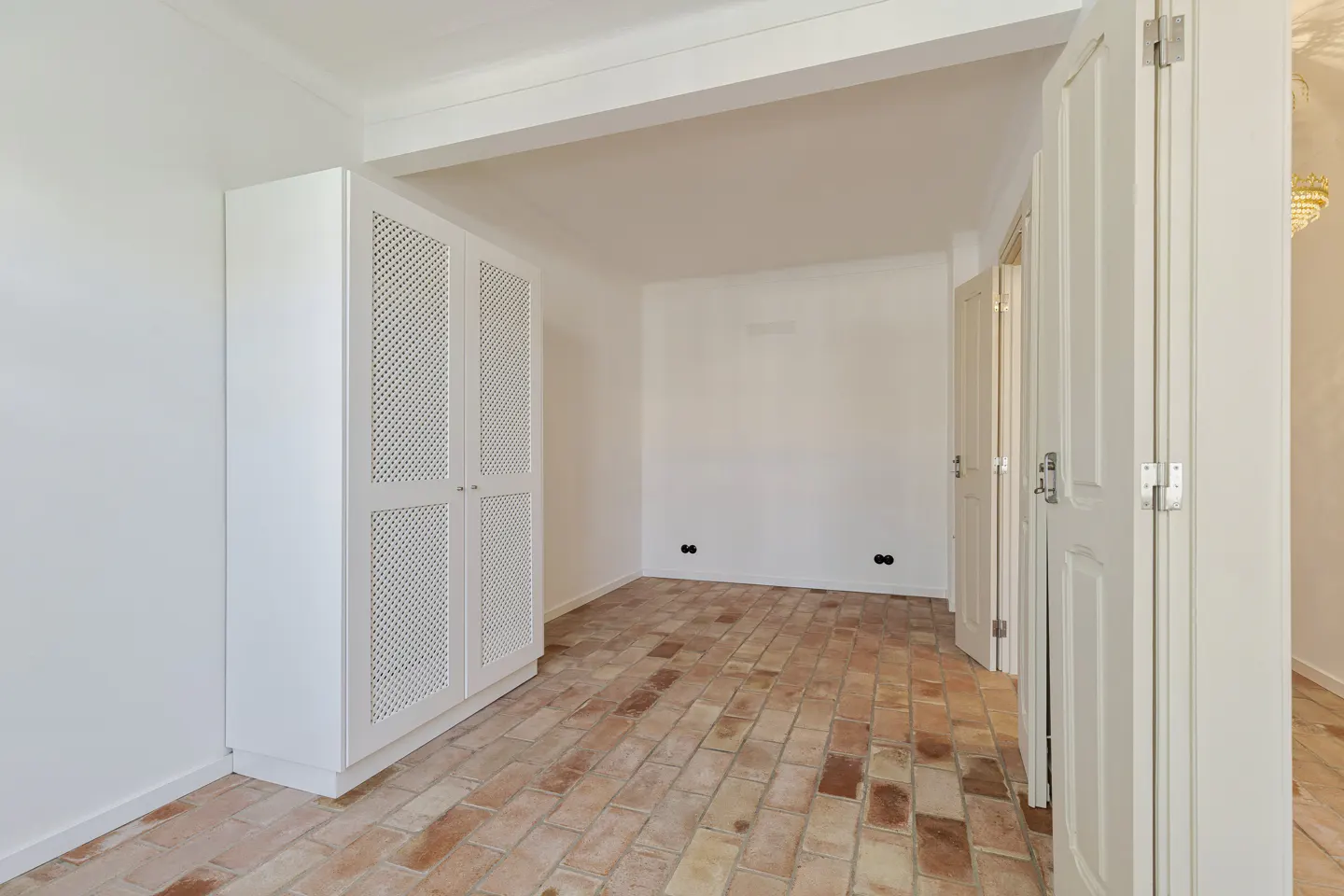 Empty room with white walls, a white wardrobe with lattice doors, and a rustic terracotta brick floor.