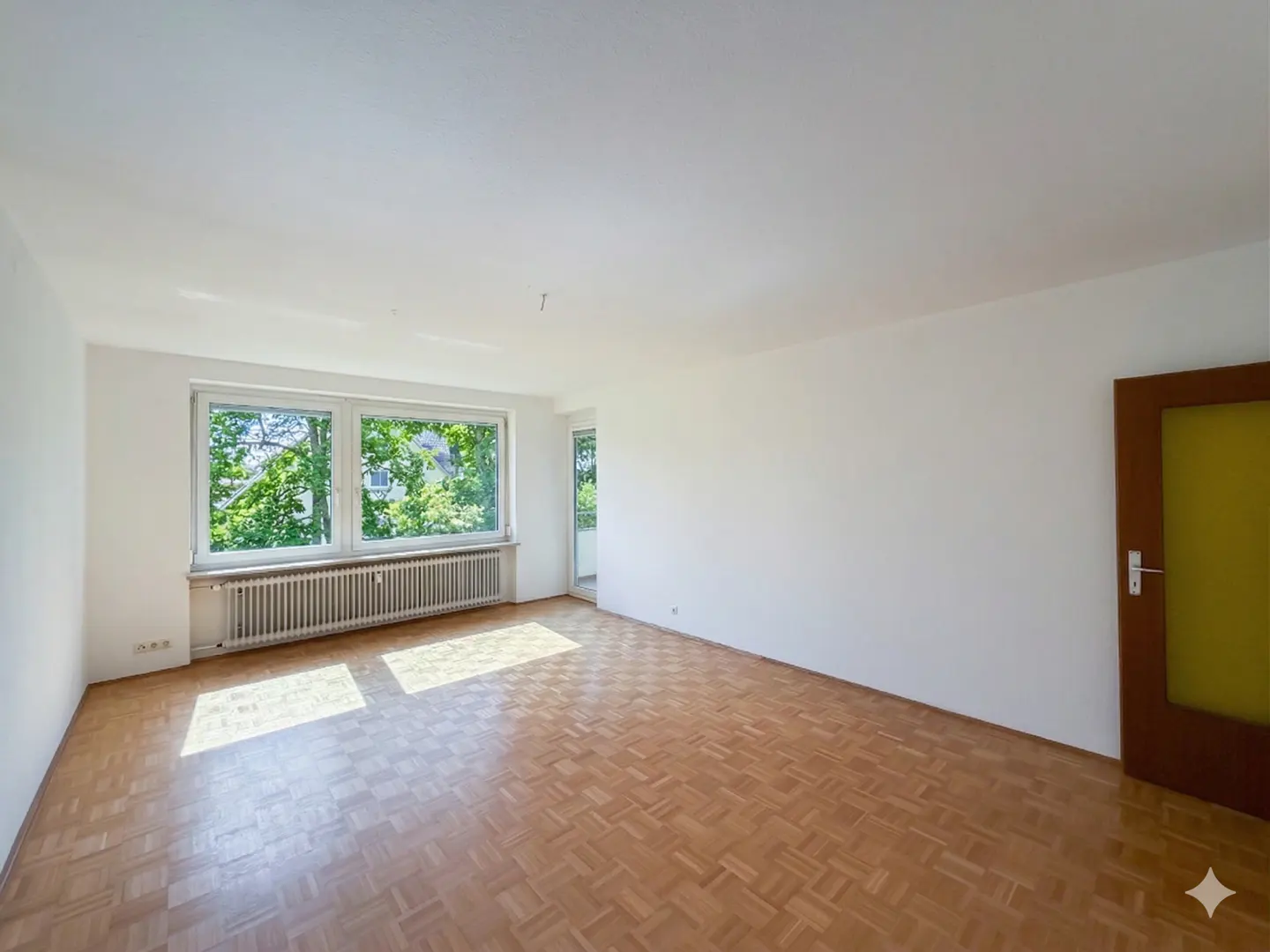 Bright, empty room with parquet floor, white walls, and a large window showing green trees outside. A radiator sits beneath the window. A brown door is on the right.