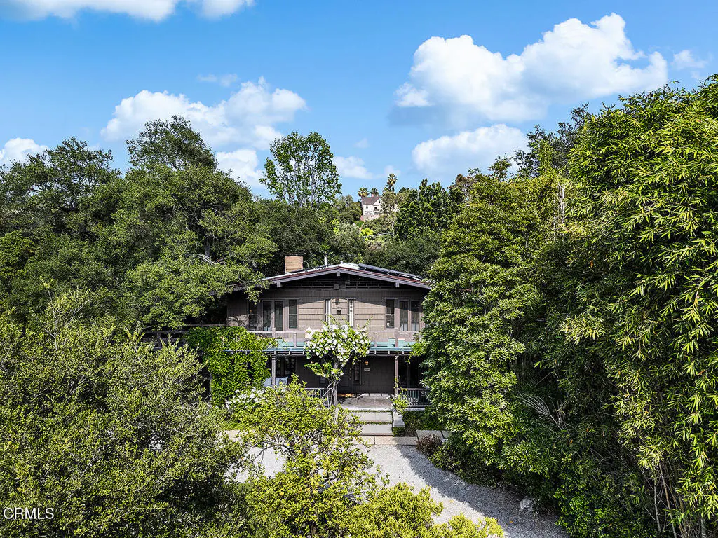 Exterior view of a two-story brown house with a chimney, surrounded by lush green trees under a blue sky with white clouds.