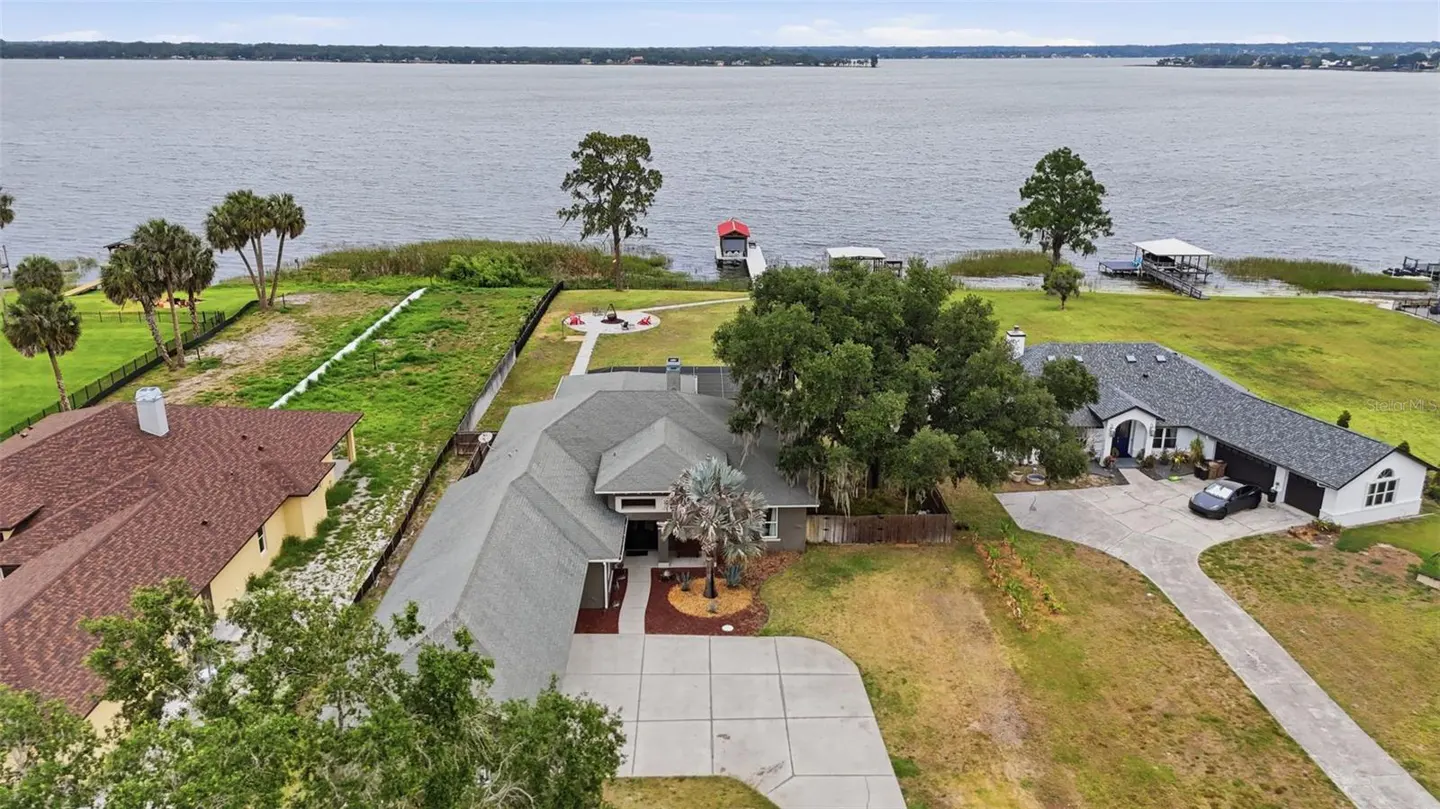 Aerial view of a gray roofed house with a long driveway, near a lake with docks and boats.