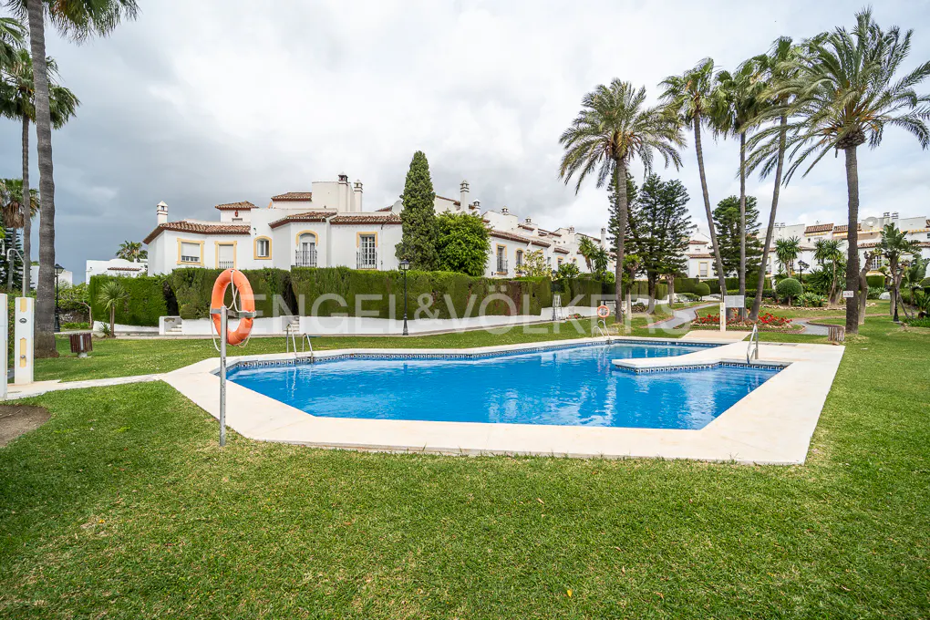 A bright blue swimming pool is surrounded by green grass and palm trees in front of white buildings.