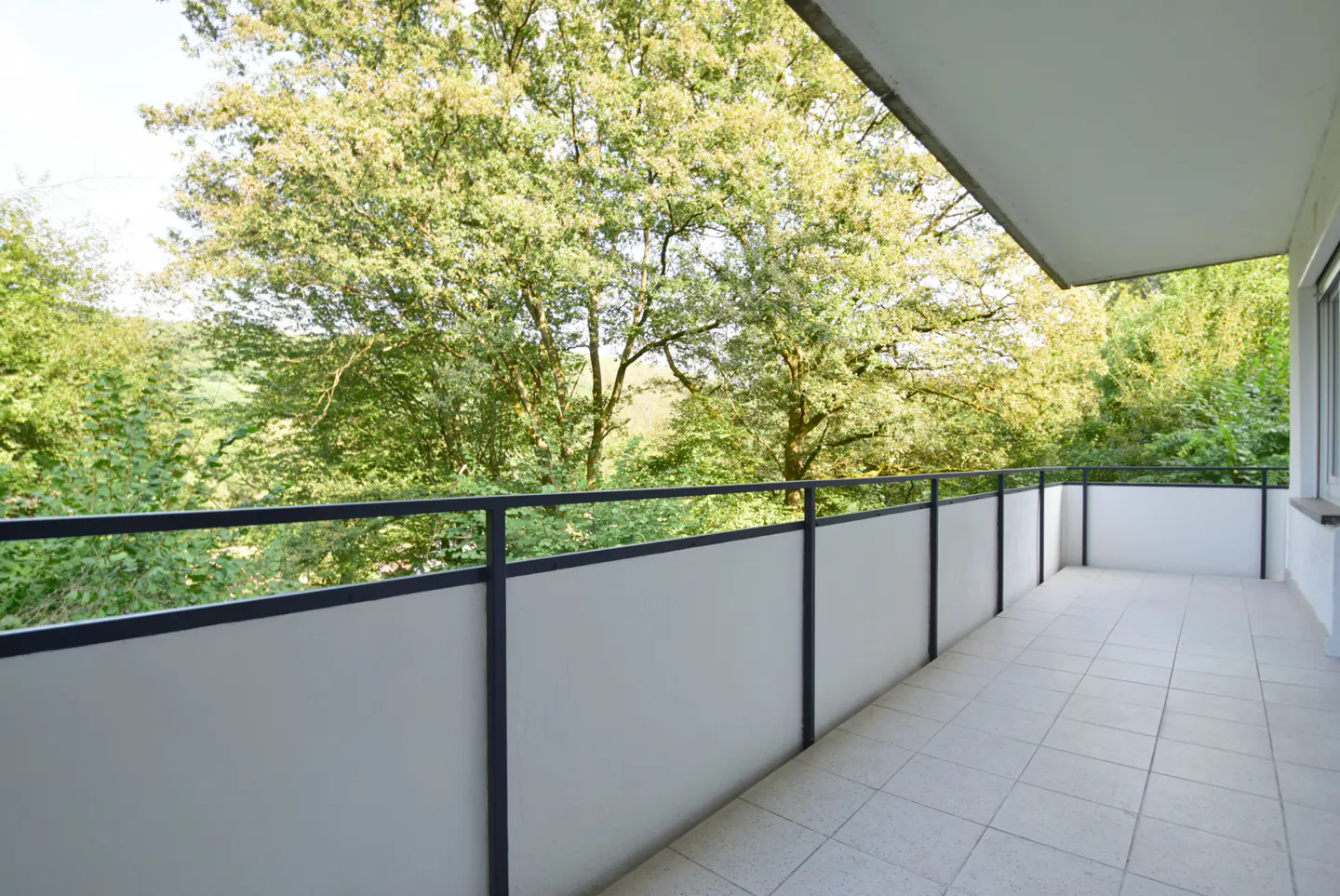 Balcony with gray tile floor, black metal railing, and frosted glass panels overlooking green trees.