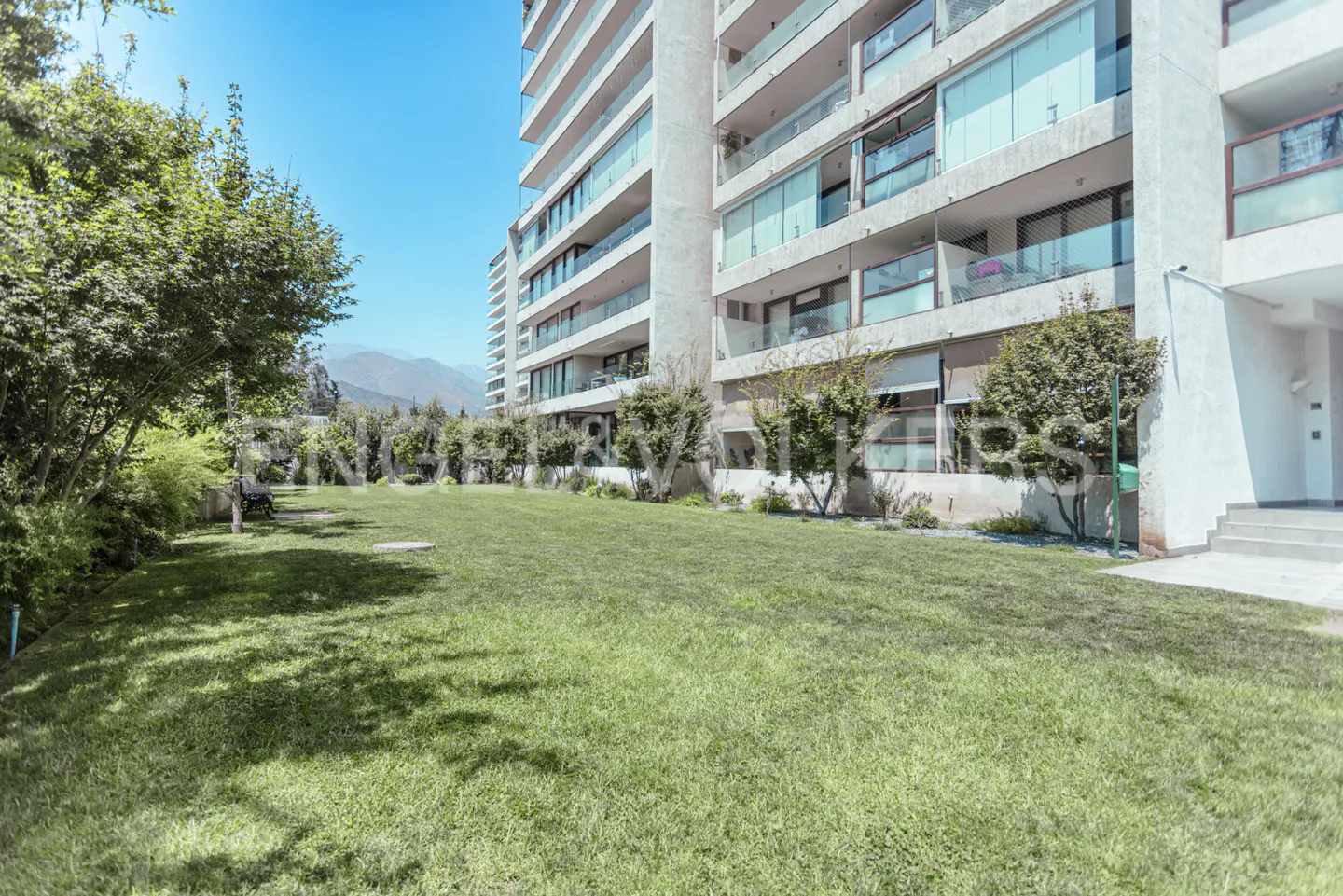 Exterior view of a modern apartment building with a green lawn and trees. Blue sky and mountains in the background.