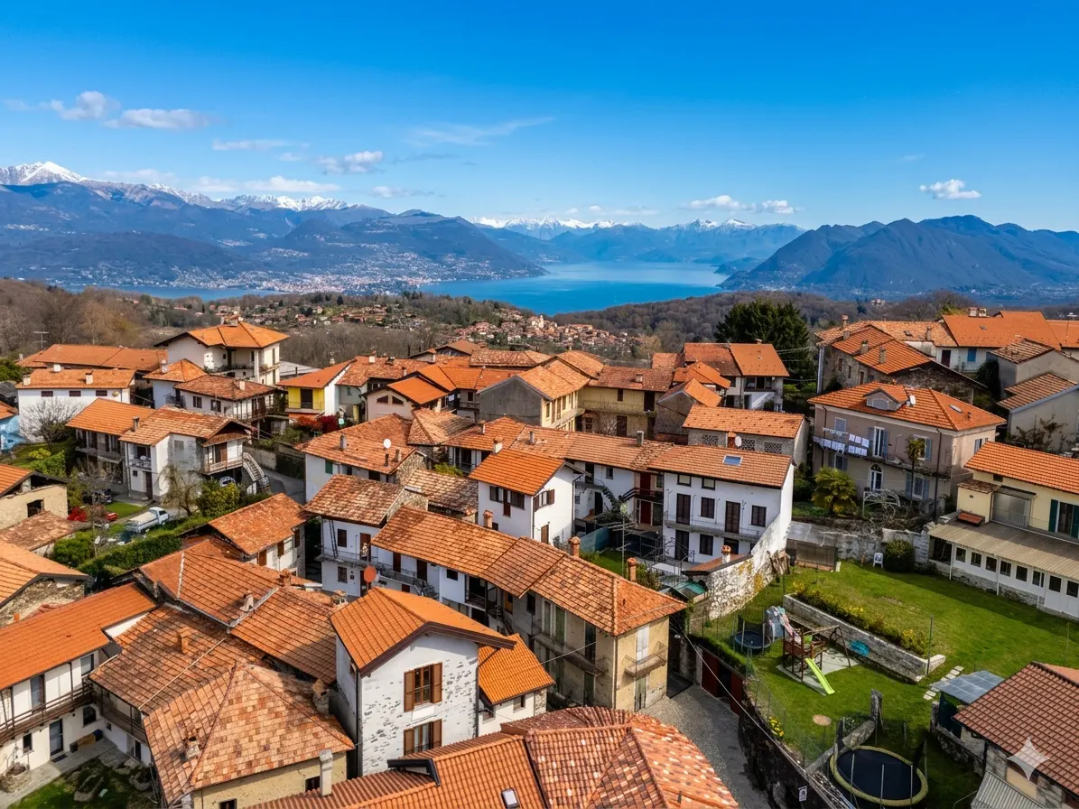 Aerial view of a village with red-tiled roofs, green lawns, and a lake with snow-capped mountains in the background.