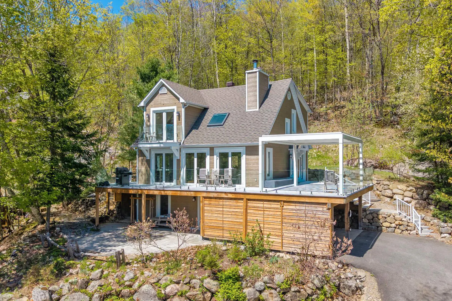 A two-story brown house with a large deck surrounded by green trees.