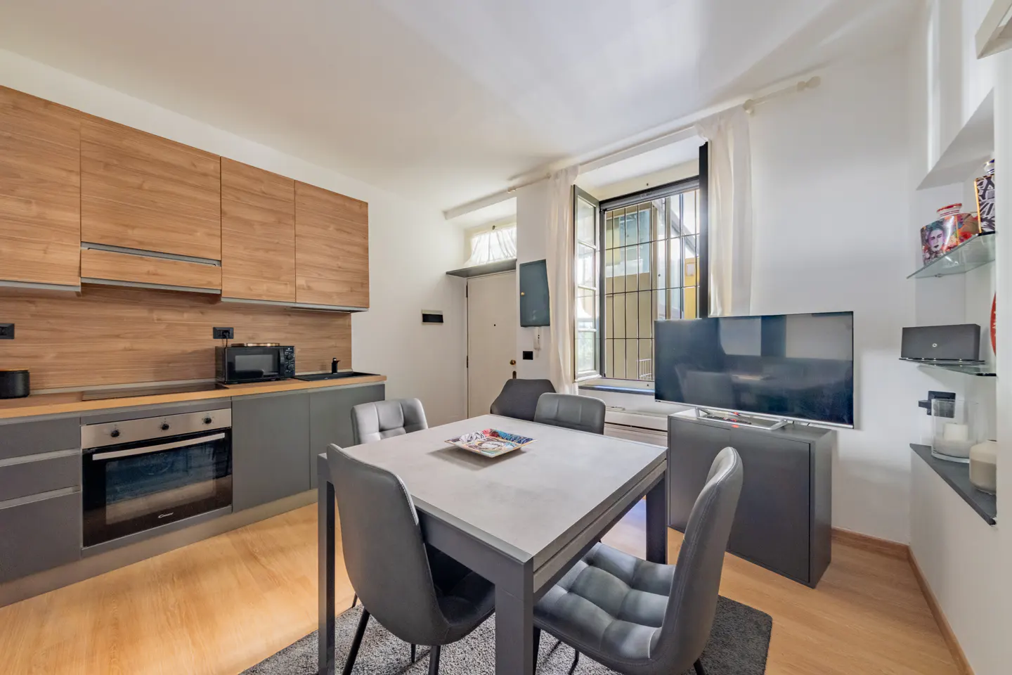Modern kitchen with wood cabinets, grey lower units, and a dining table with four grey chairs near a window and TV.