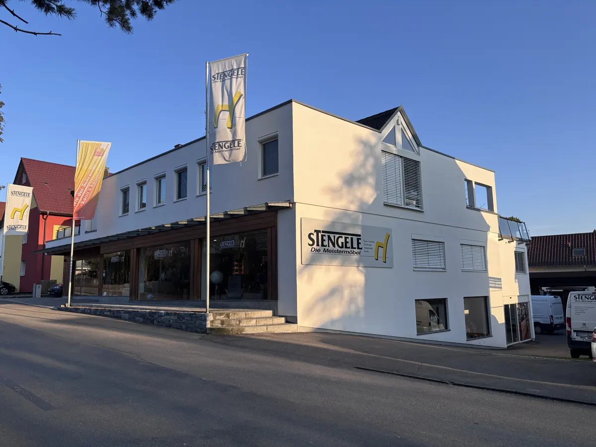 Exterior view of Stengele furniture store, a modern white building with large display windows and flags bearing the company logo.