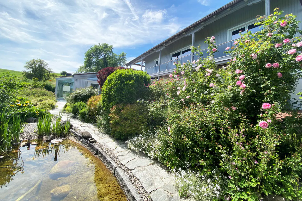 A gray house with a balcony is surrounded by lush gardens with pink roses and a pond with clear water.