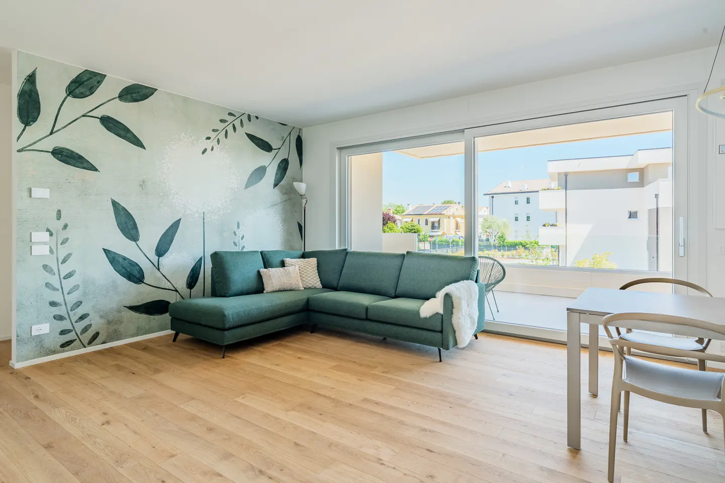 Bright living room with a green sectional sofa, floral wallpaper, and a dining table near a large window.