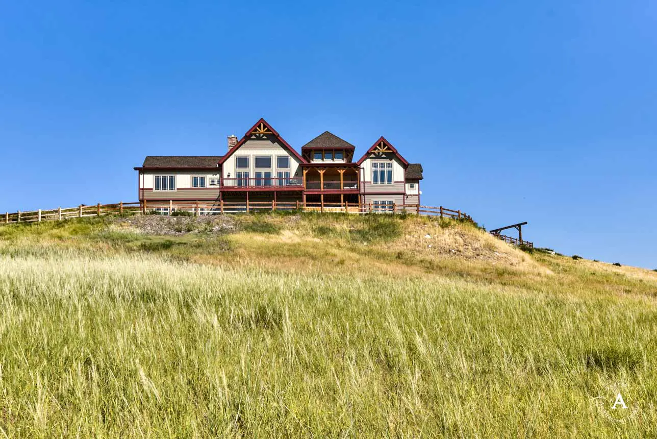 A large, two-story house sits atop a grassy hill under a clear blue sky. A wooden fence surrounds the property. Tall grass fills the foreground.