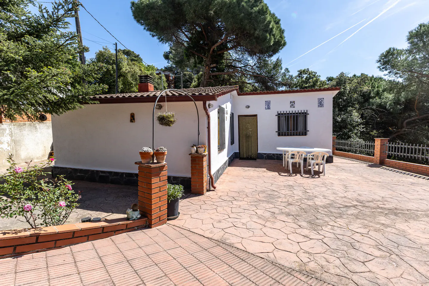 Exterior view of a white house with a red tile roof and brick accents, surrounded by greenery and a patterned stone patio.