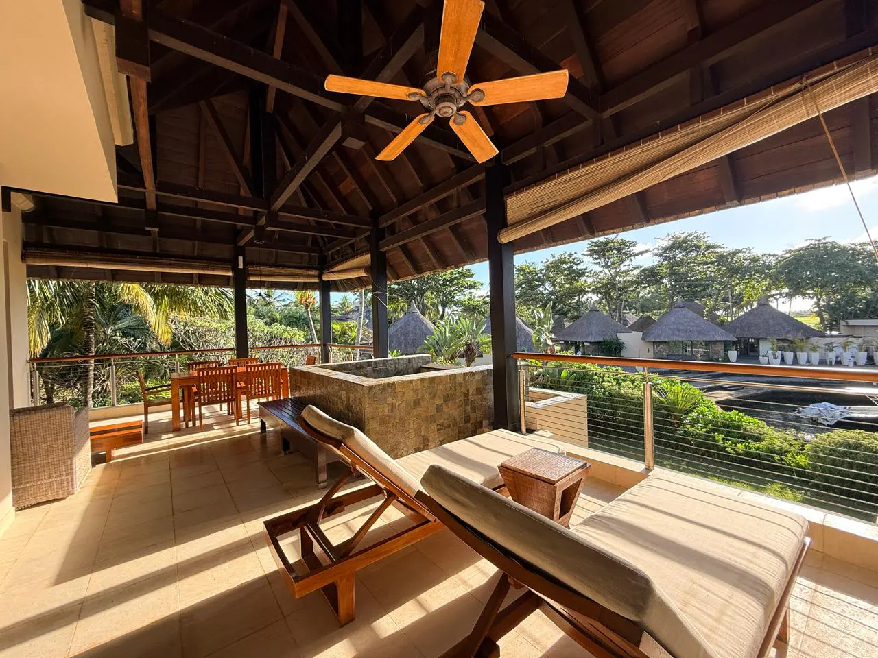 Covered patio with two lounge chairs, a table and chairs, and a ceiling fan. Outside is a pool and thatched roof huts.