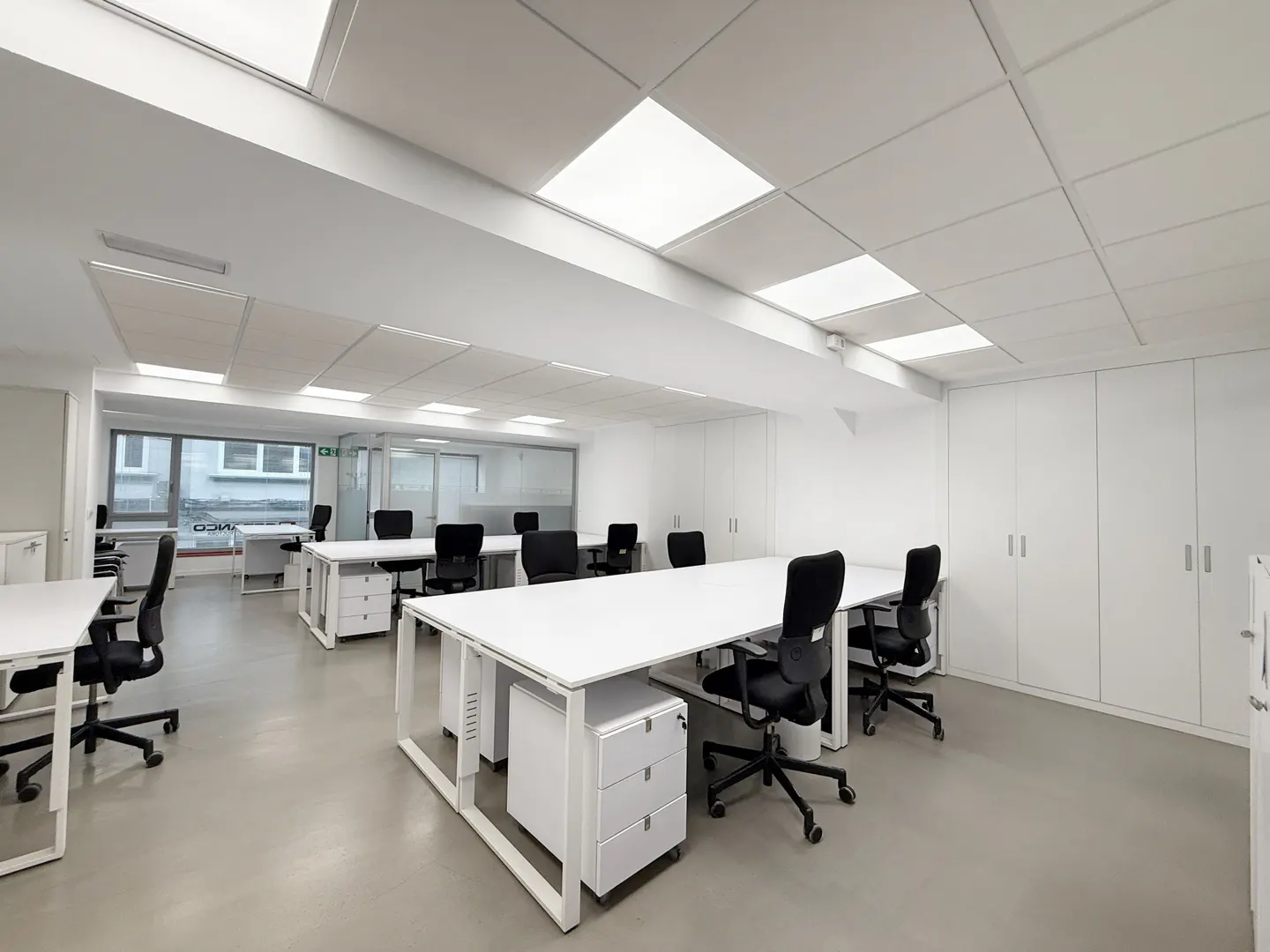 Modern office space with white desks, black ergonomic chairs, white storage cabinets, and bright recessed ceiling lighting.