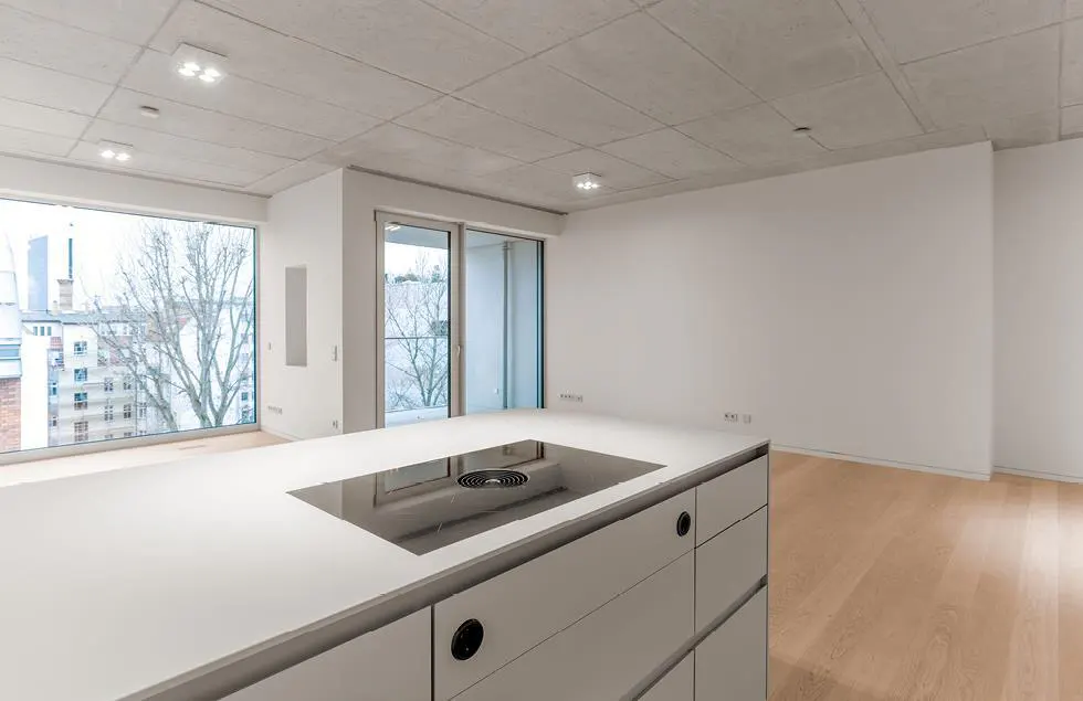 A modern, empty apartment with a white kitchen island, light wood floors, and large windows overlooking a cityscape.