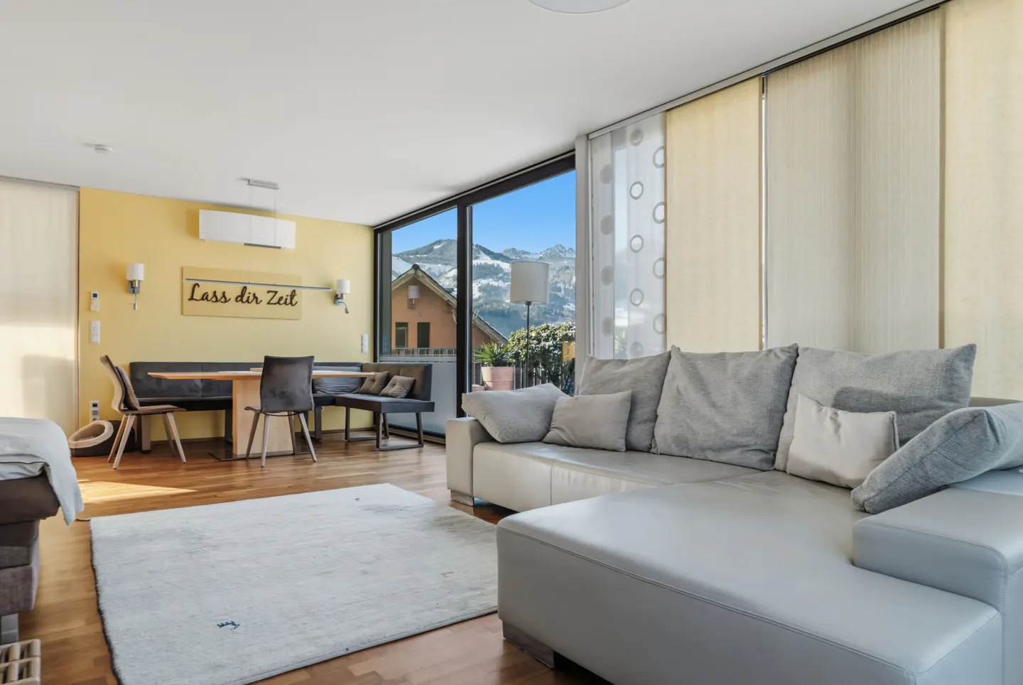 Bright living room with a gray sectional sofa, dining table, and a view of snow-capped mountains through a large window.