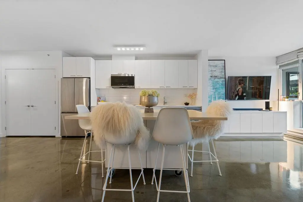 A modern, all-white kitchen with a central island and four bar stools, two with furry white covers. Stainless steel appliances and polished concrete floors.