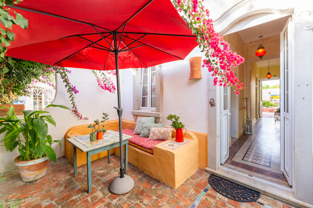 Outdoor patio with red umbrella, brick floor, and built-in seating. Pink bougainvillea climbs the white walls. Open doorway leads to a hallway.