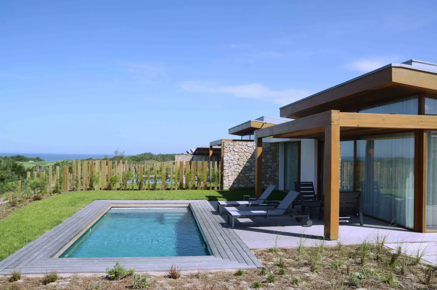 Modern home exterior with a small pool, lounge chairs, and a wooden pergola. Ocean view in the background.