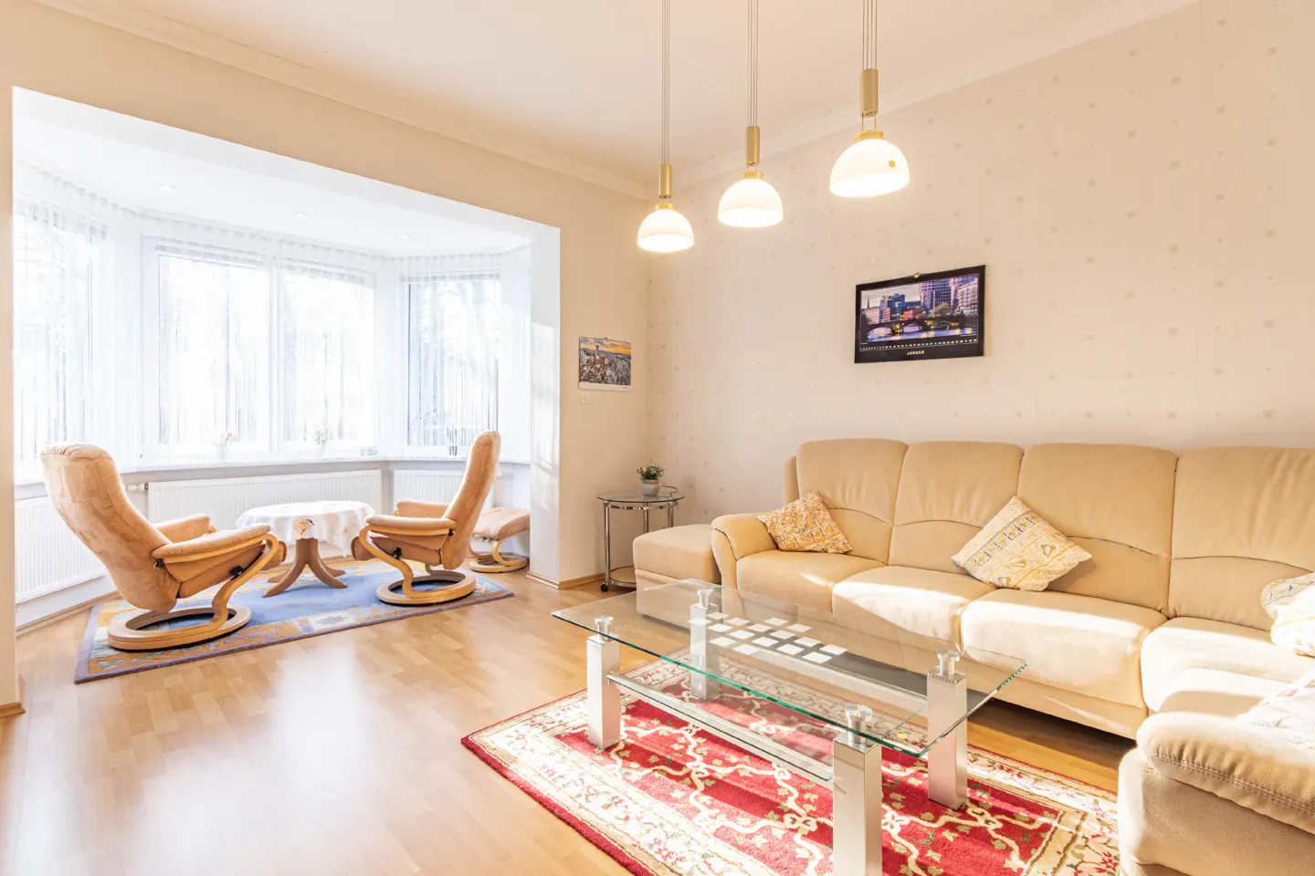 A bright living room with a beige sofa, glass table, and two tan armchairs in a bay window. Three pendant lights hang above.