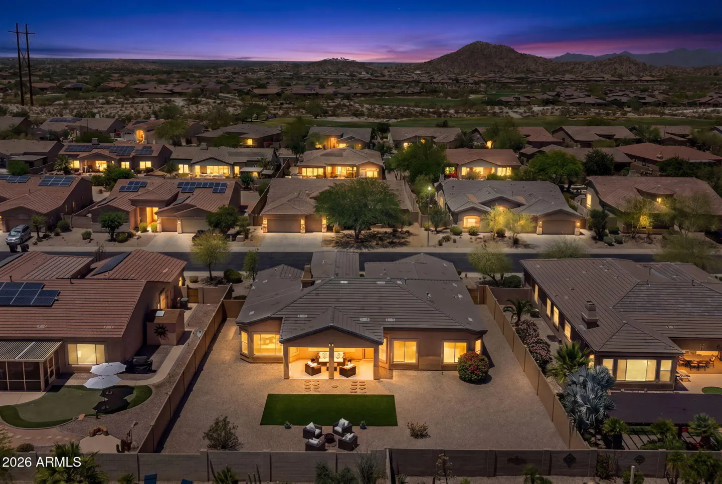 Aerial view of a desert neighborhood at dusk, with warm lights glowing from the houses and mountains in the distance.