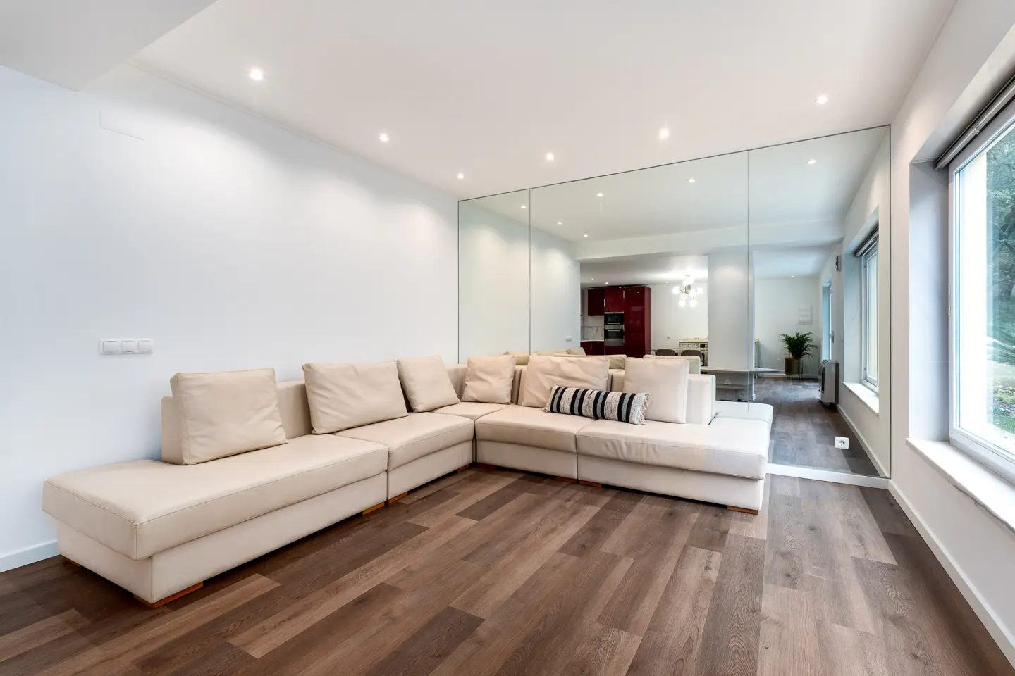 Bright living room with a large beige sectional sofa, wood floors, and a wall mirror reflecting the kitchen.