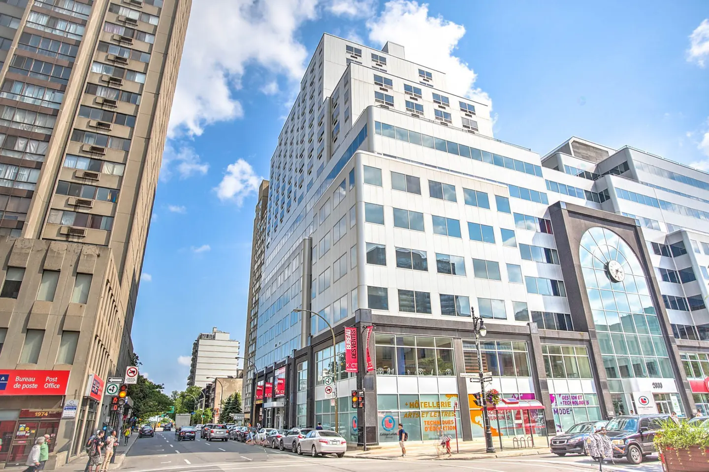 Low-angle view of a modern white office building with large windows on a sunny city street with cars and pedestrians.