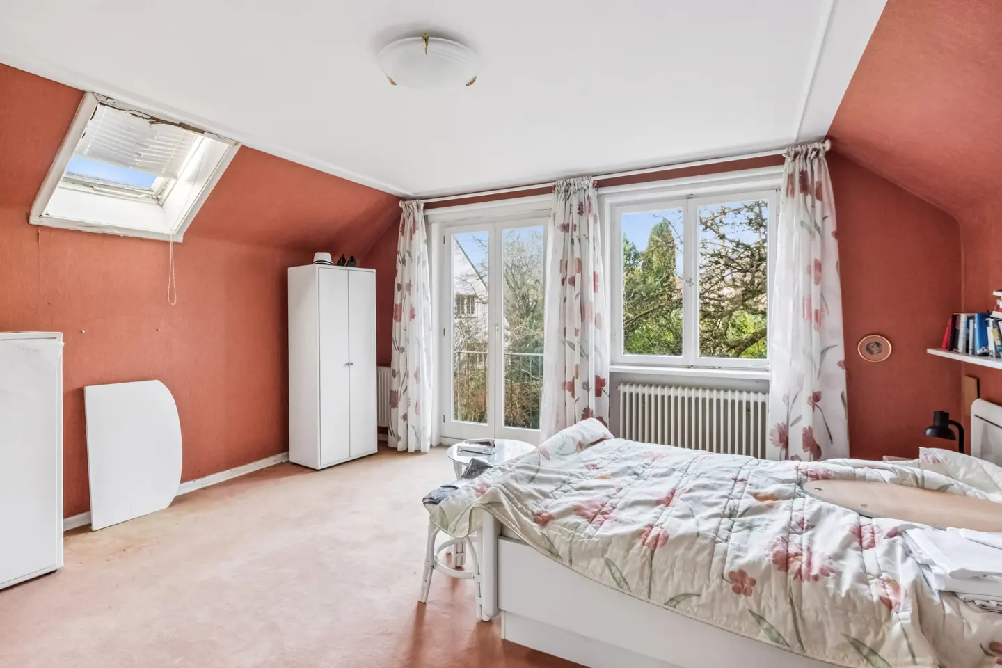 Attic bedroom with red walls, white trim, and floral bedding. Windows with curtains overlook trees. A skylight and white wardrobe are also visible.