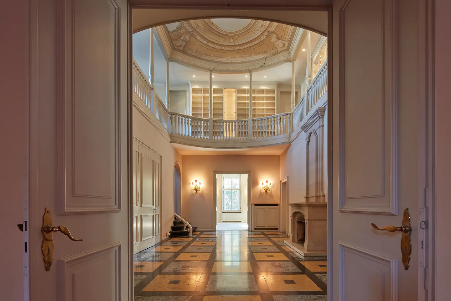 View through open white doors into a grand foyer with a balcony, bookshelves, and patterned floor.