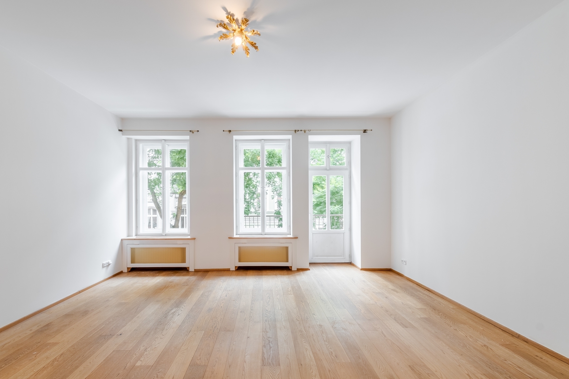 Empty room with white walls, light wood floors, three windows, and a decorative gold ceiling light fixture. [AI-generated text]