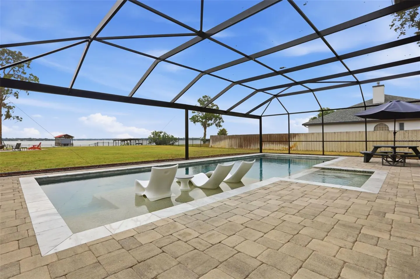 Screened-in pool with three white lounge chairs in the water, a hot tub, and a picnic table on a brick patio. A lake is visible in the background.
