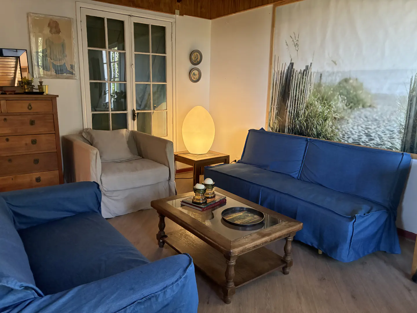 Living room with blue sofas, a beige armchair, and a wooden coffee table. A beach scene print hangs above the sofa.