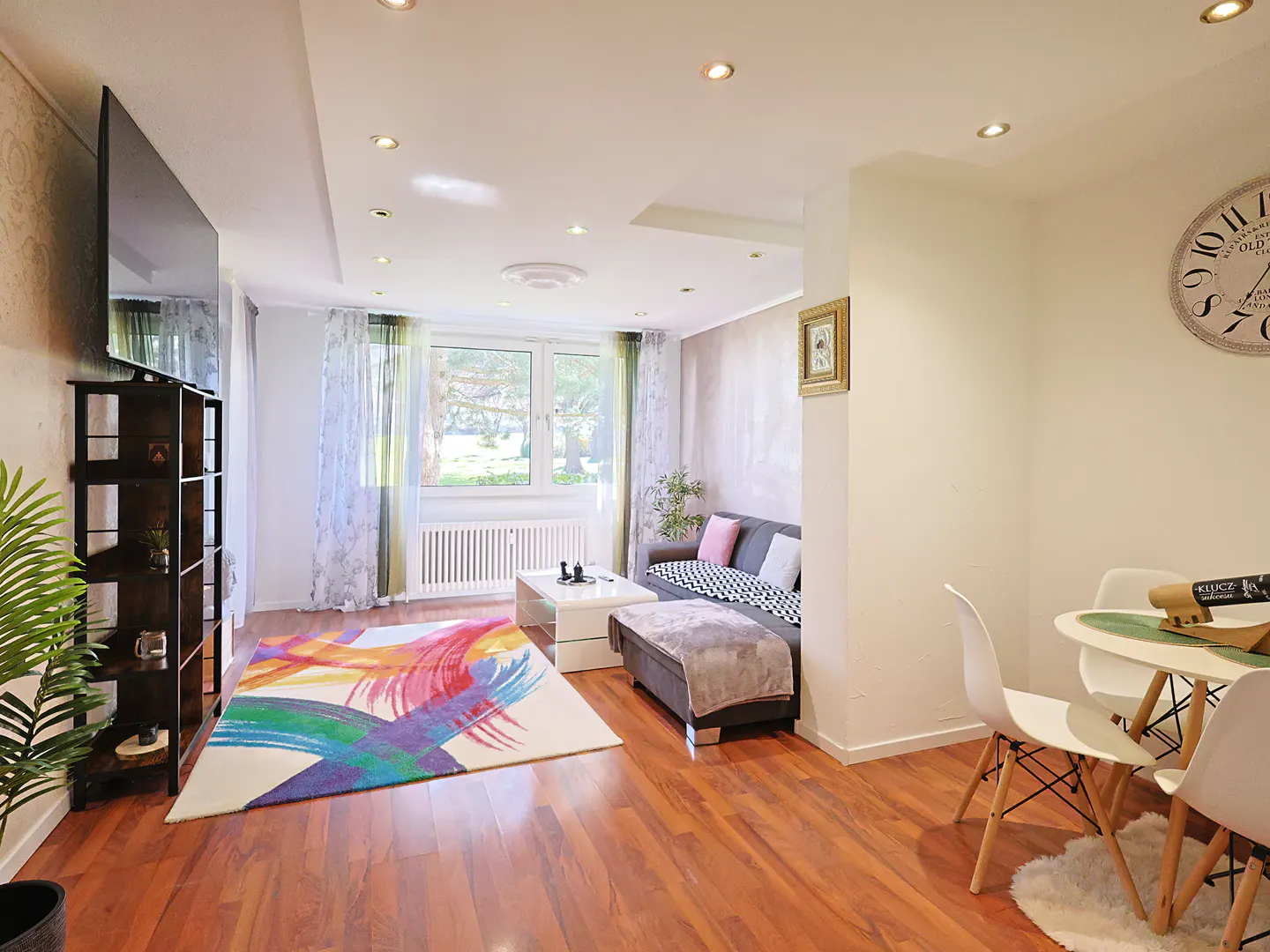 Bright living room with hardwood floors, a colorful rug, a gray sofa, and a white dining table with chairs.