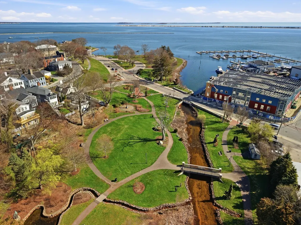 Aerial view of a park with a stream, bridge, and harbor with boats in the background. Houses and trees surround the park.