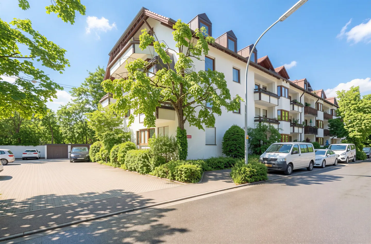 White multi-story apartment building with balconies, surrounded by lush green trees and a paved parking lot with cars.