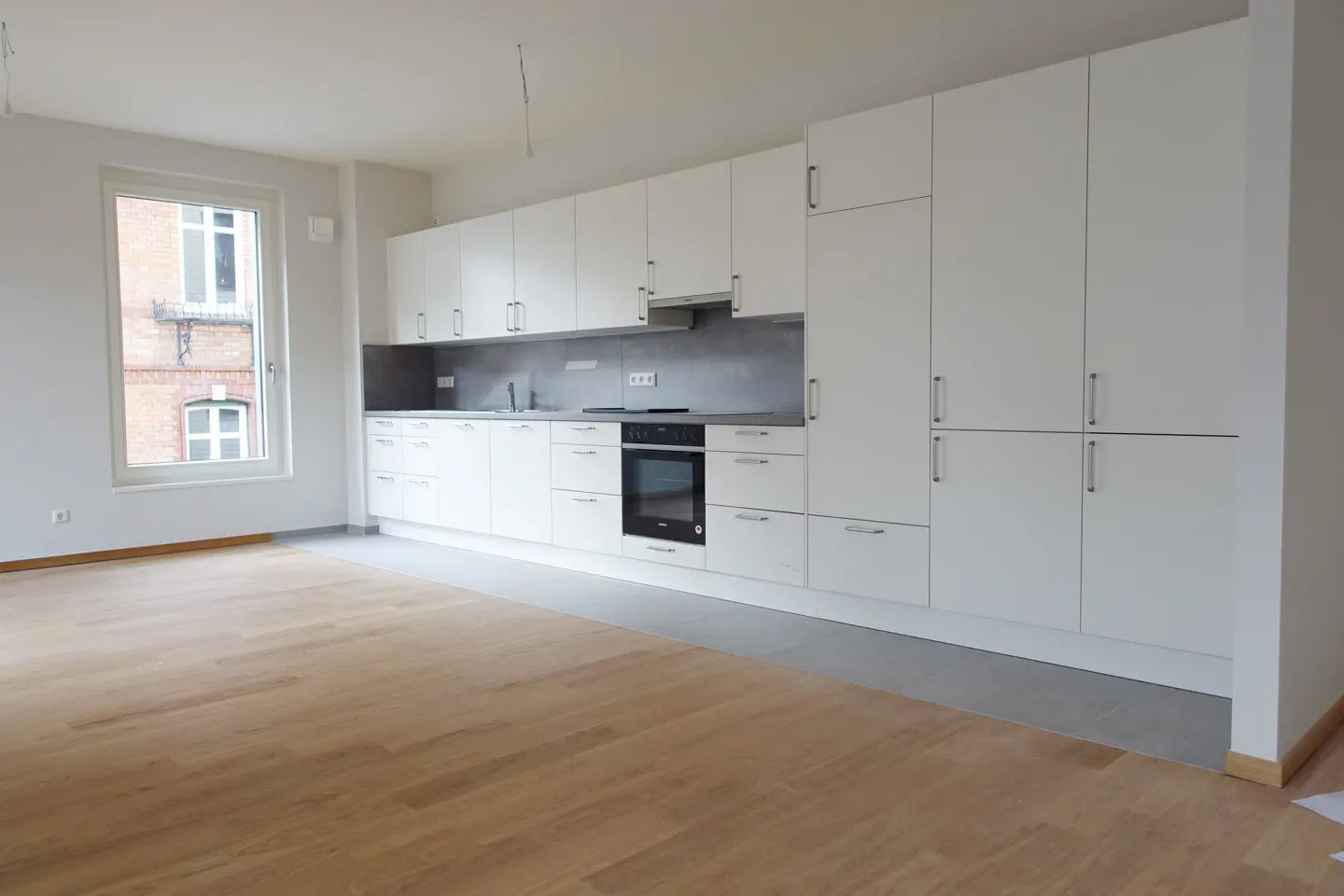 A bright, modern kitchen with white cabinets, gray countertops, and light wood floors. A window shows a brick building outside.